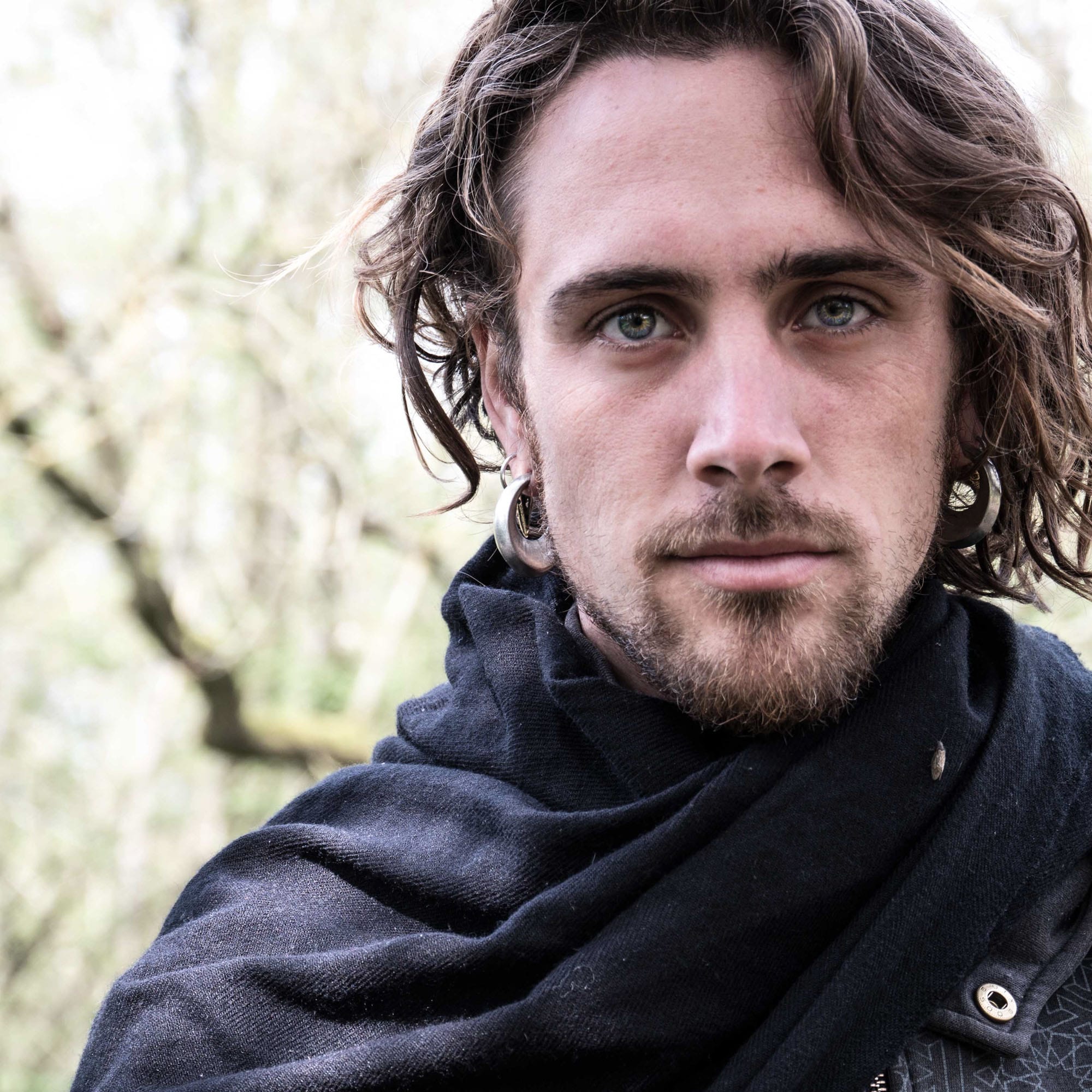 A man with medium-length wavy hair and a beard is looking directly at the camera. He is wearing a dark scarf wrapped around his shoulders and has SAN Minimalist Diamond Ear Weights in Gold (2 gauge) by Kaya Movement in both ears. The background is blurry, showing what seems to be a natural, outdoor setting with trees.